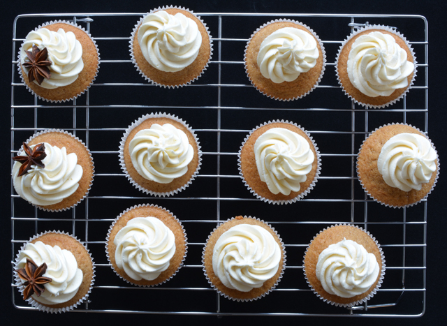 top view image of a cooling rack with a freshly baked chai tea spiced cake topped with buttercream and three cupcakes adorned with star anise