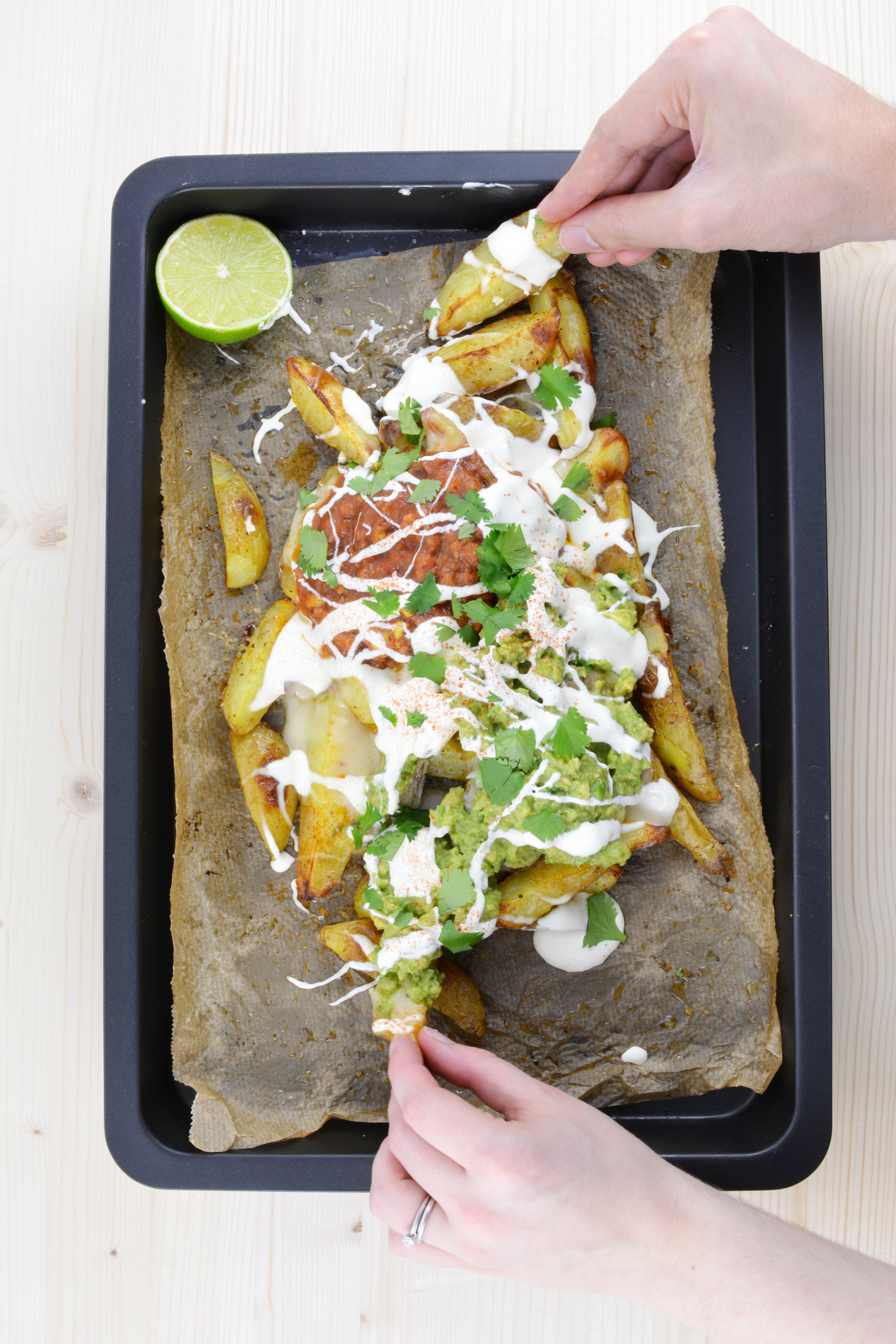 an overhead view of a baking tray featuring potato wedge nachos generously topped with guacamole, salsa, and sour cream. Two hands are reaching for a potato wedge on the tray