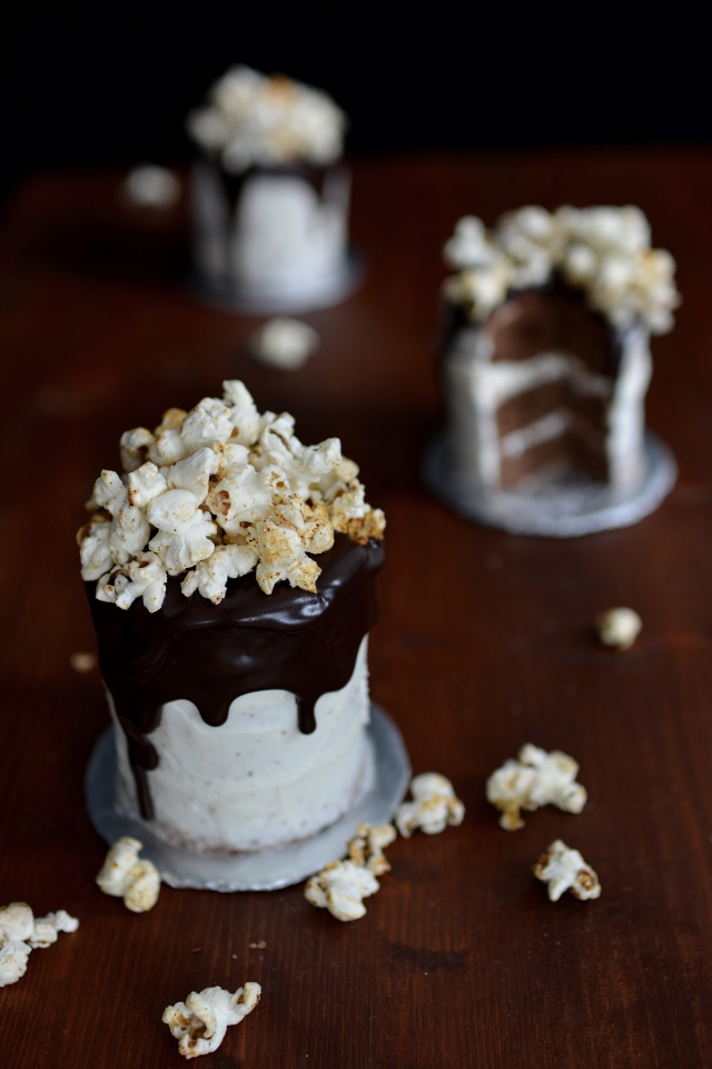 top view of three chocolate cakes with chocolate syrup drizzle and popcorn toppings. one cake in the foreground with scattered popcorn, and one cake in the background is partially sliced