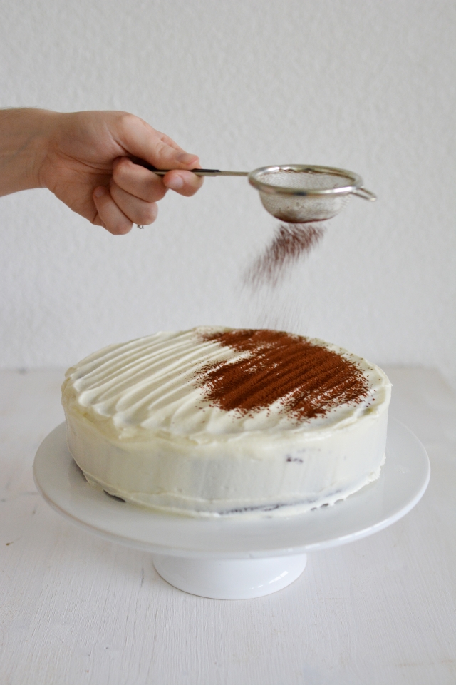 image of a hand using a strainer to sprinkle cocoa powder onto a chocolate cake covered with cream cheese frosting