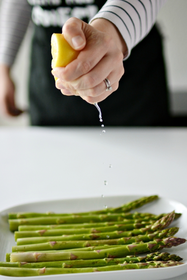 a hand squeezing a fresh lemon over a bunch of chargrilled asparagus stems