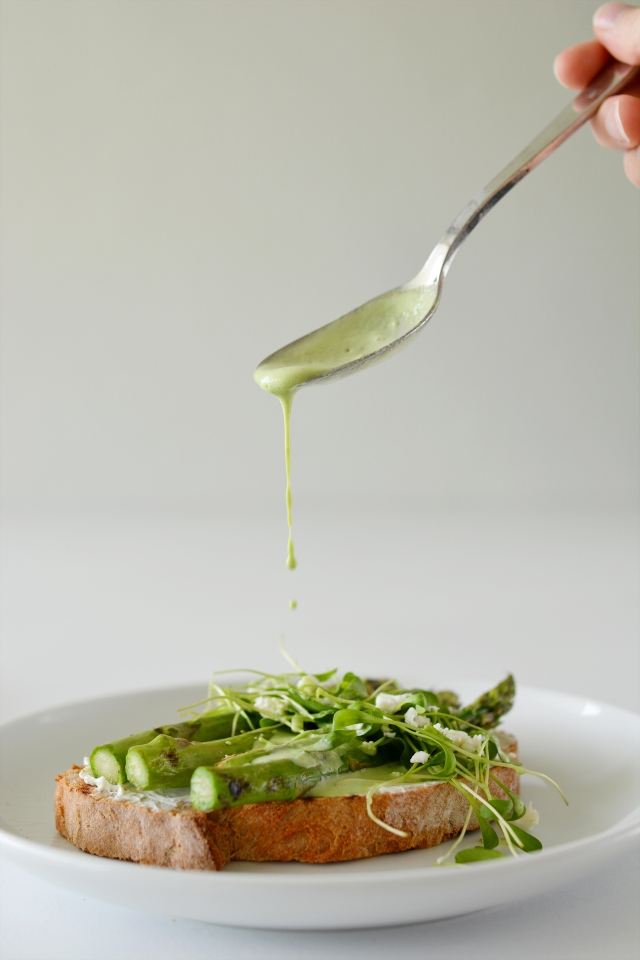 a spoon drizzling yogurt dressing over toasted bread with asparagus, parsley, cress, and feta cheese