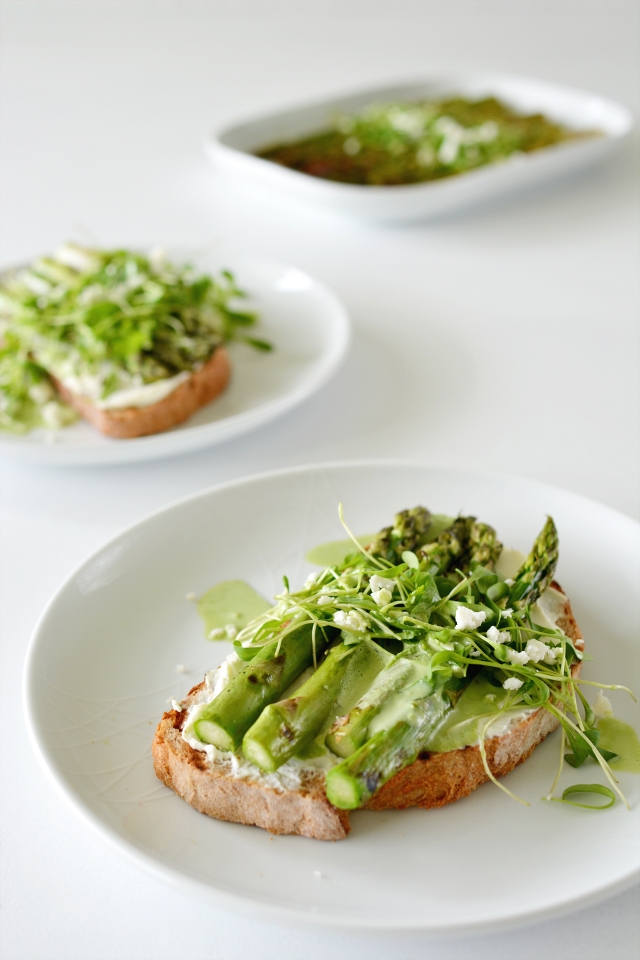 close-up image of a single slice of toasted bread featuring grilled asparagus on top. The dish is beautifully adorned with a drizzle of yogurt dressing, fresh cress, and crumbled feta cheese