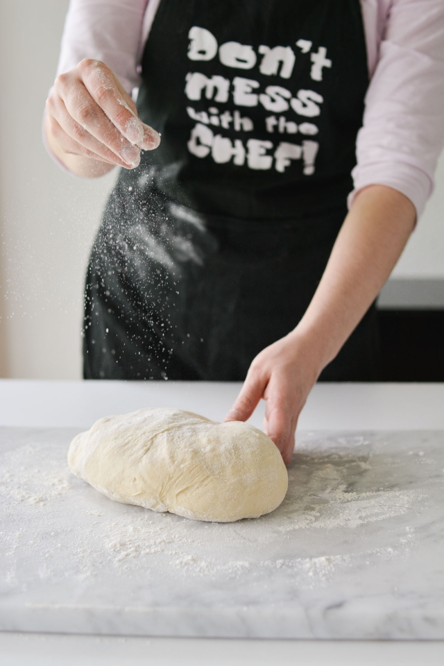 woman spreading flour onto a kneaded dough on a well-floured surface. Her hands are engaged in the process, creating a rustic and tactile scene of dough preparation