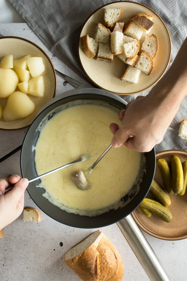 two hands dipping a cut of bread into the swiss cheese fondue mixture