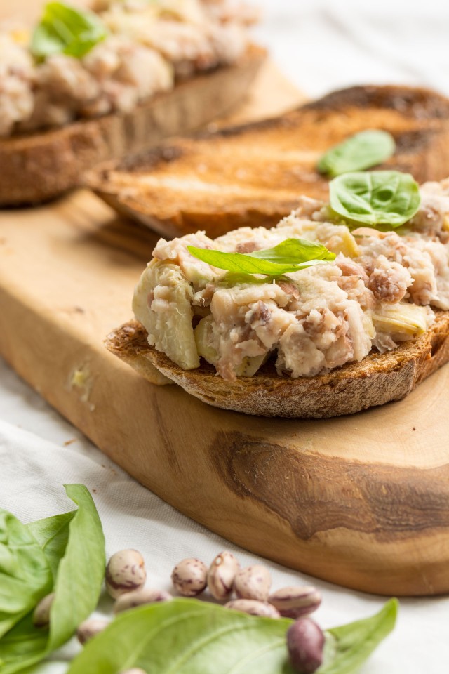 close up image of a crusty bread topped with a flavorful mix of two beans and artichoke on a cutting board with fresh basil leaves