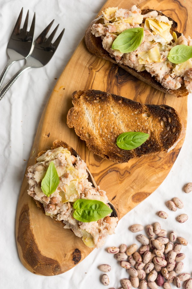 top down image of a crusty bread topped with a flavorful mix of two beans and artichoke on a cutting board with fresh basil leaves