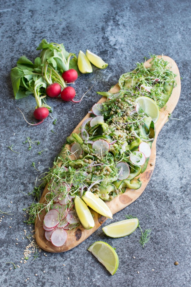 Top view of a cucumber and lentil salad with radishes on a wooden board and a bunch of radishes and lemon slices on the side. 