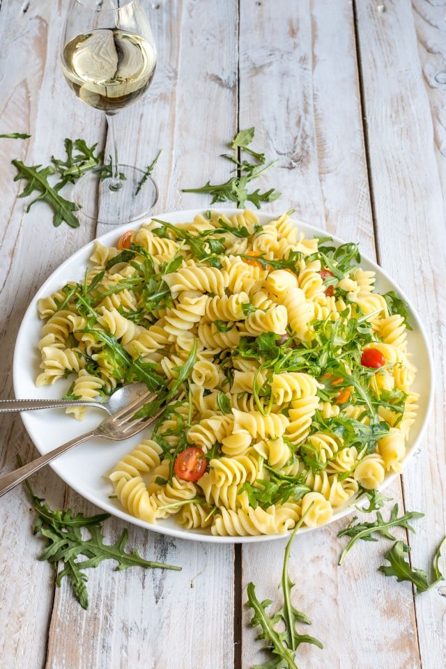 Lemon Arugula Pasta Salad on a white plate on a white wooden table with a glass of white wine.