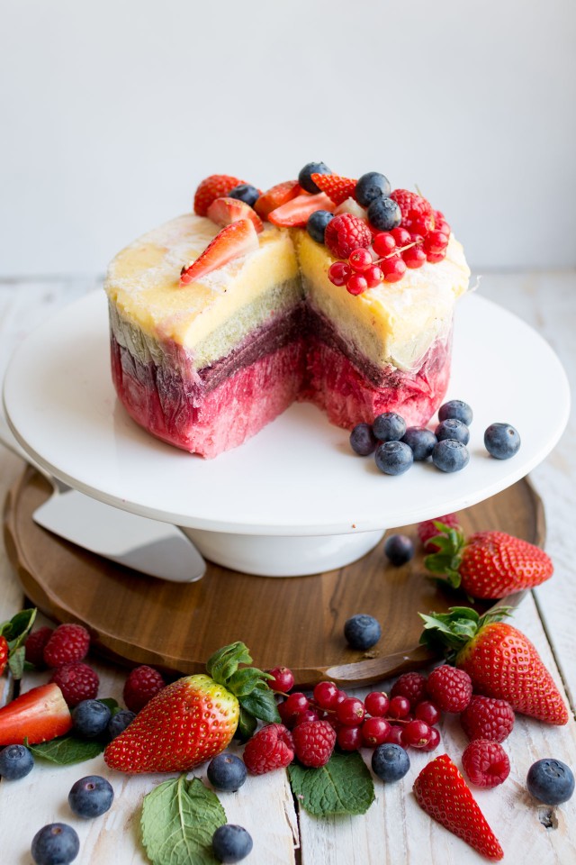 an image of a delicious and colorful yogurt layer cake in a pedestal with a slice, topped with strawberries, rapsberries, and blueberries