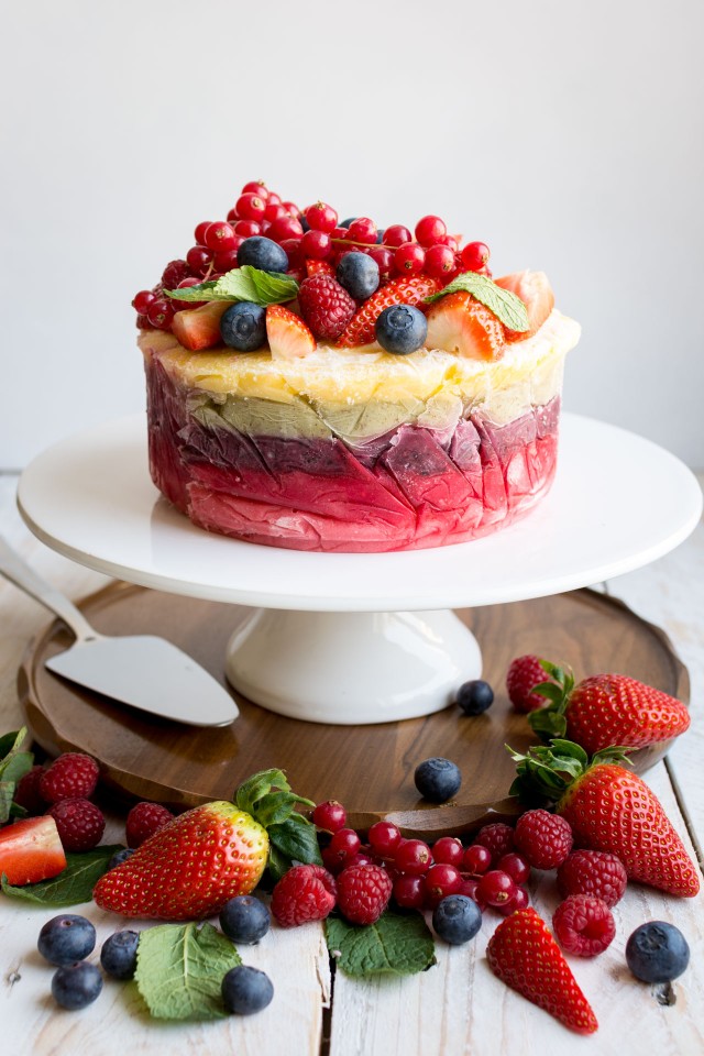 front image of a delicious and colorful yogurt layer cake in a pedestal topped with strawberries, rapsberries, and blueberries